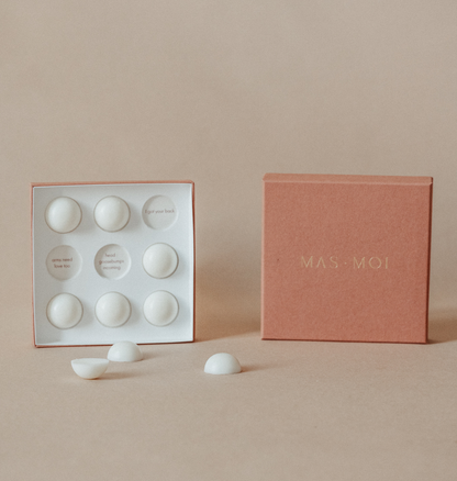 An open masmoi box revealing floral scent massage oil pearls inside, each designed to melt into nourishing oil for a soothing massage. The inner packaging includes playful text such as 'I got your back' and 'head goosebumps incoming,' adding a personalised touch. Set against a warm beige background, this luxurious product is perfect for a massage workshop or self-care ritual.