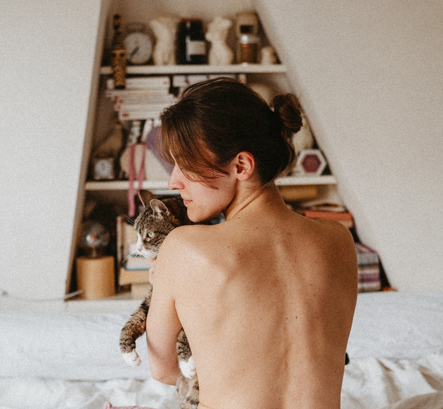 A topless woman sits on a bed, holding a cat, her back exposed and ready to be massaged during a massage workshop.