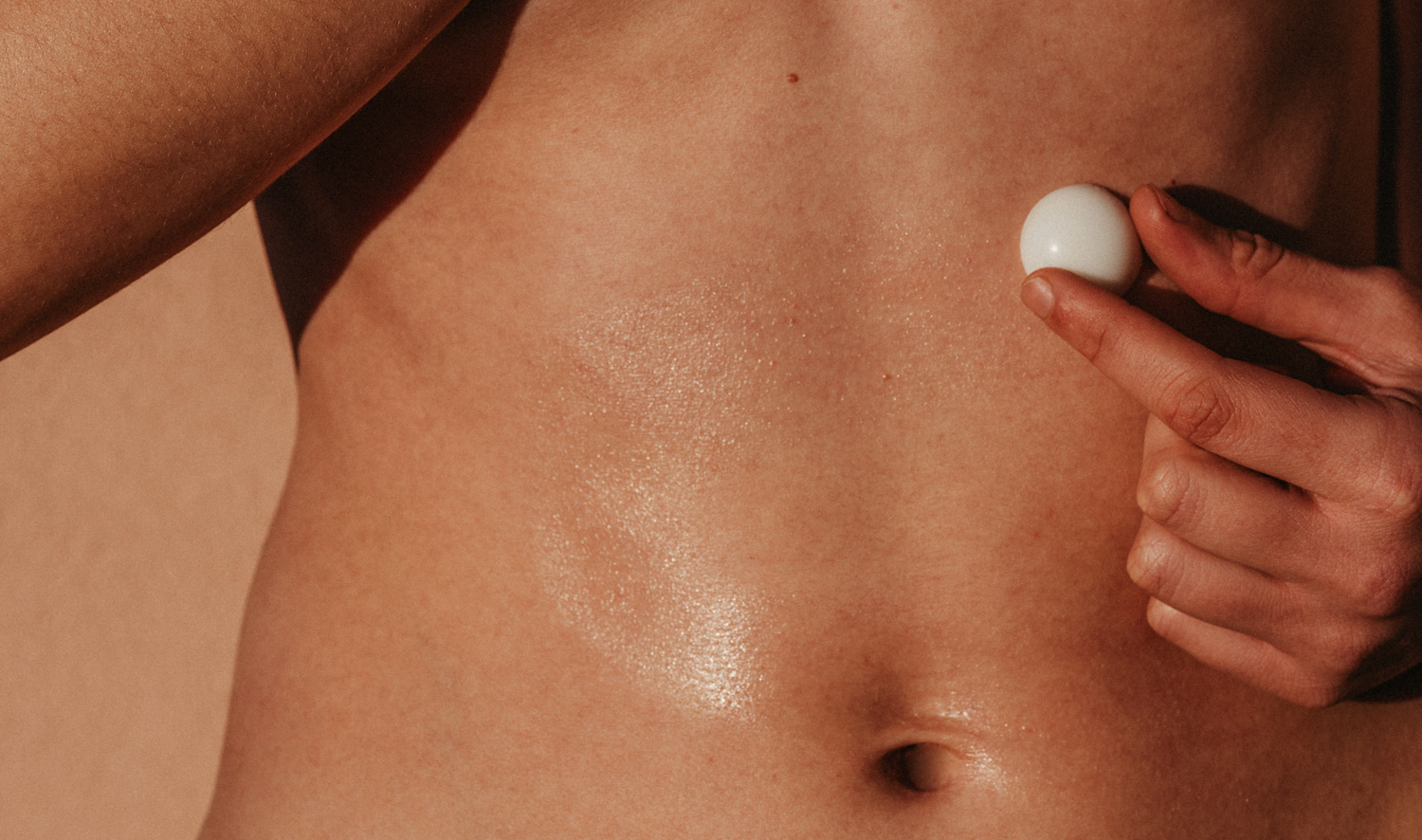 A close-up of a person’s bare torso during a massage workshop, holding a lavender massage oil pearl against their skin. Their fingers gently press the pearl, which is designed to melt upon contact, releasing nourishing oils. The warm lighting and dewy skin enhance the intimate and therapeutic feel of the massage experience.