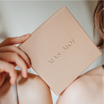 A close-up of two people, their shoulders touching, holding a box of masmoi massage pearls, in gold lettering. The soft lighting and neutral tones create a luxurious, intimate feel, suggesting a high-end product for a massage experience.