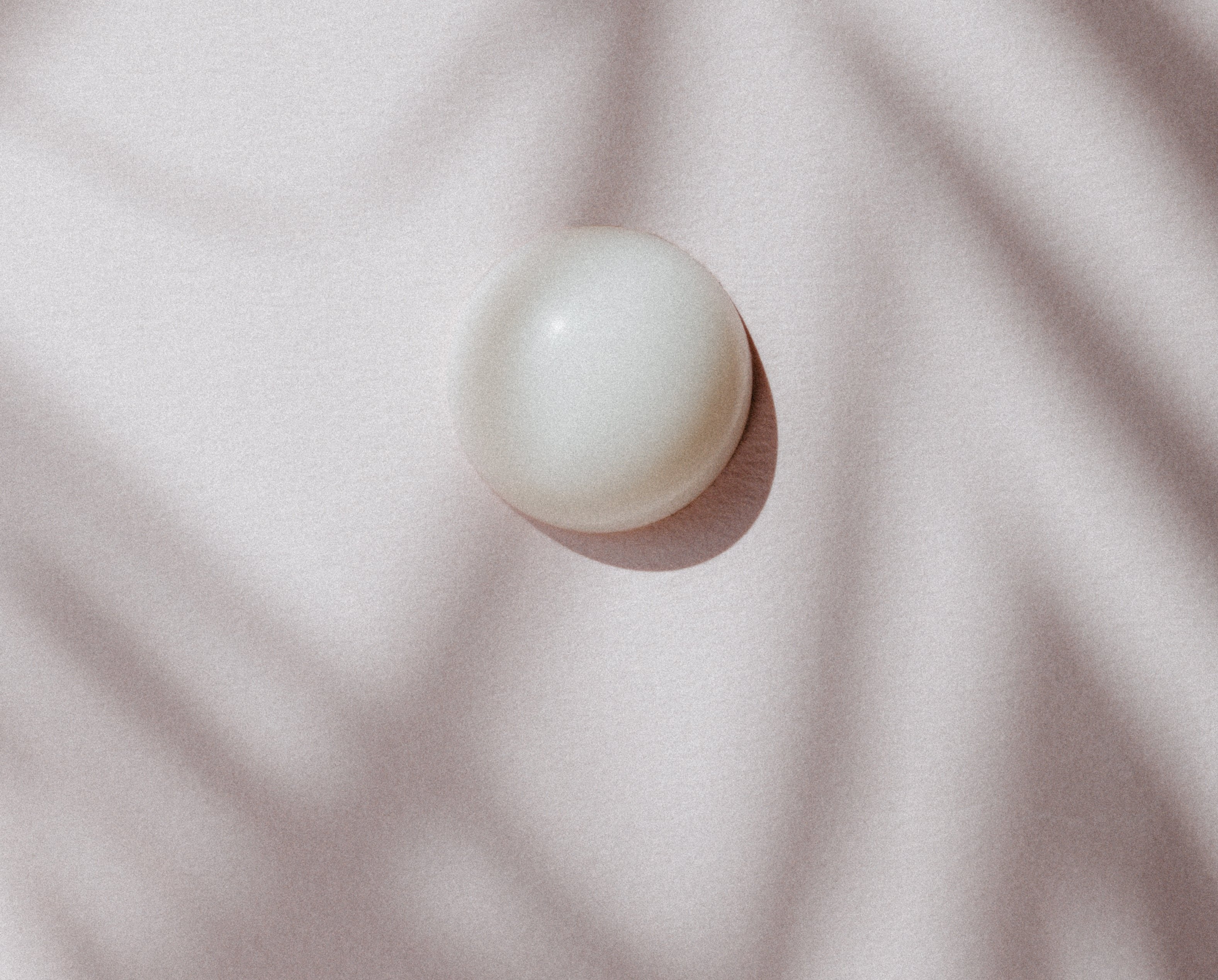 A single massage oil pearl rests on a soft, neutral fabric, casting a gentle shadow in the sunlight. This minimalist composition highlights the natural skincare ingredients, making it a perfect product for a massage experience.